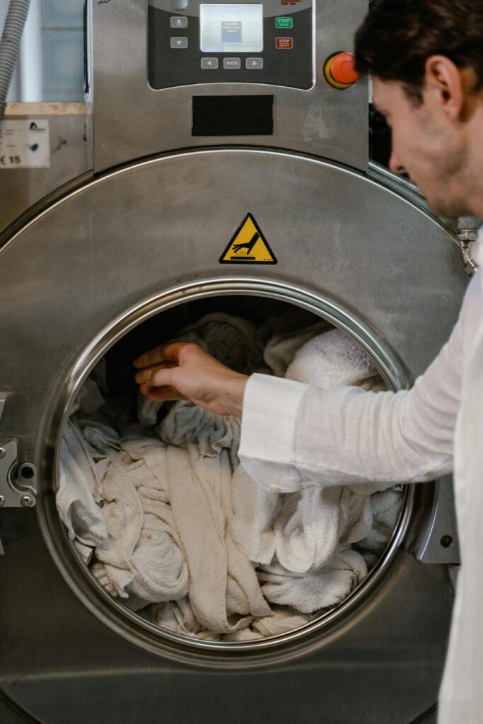 Man in white shirt loading laundry into a large industrial washing machine, focusing on hand and towel.
